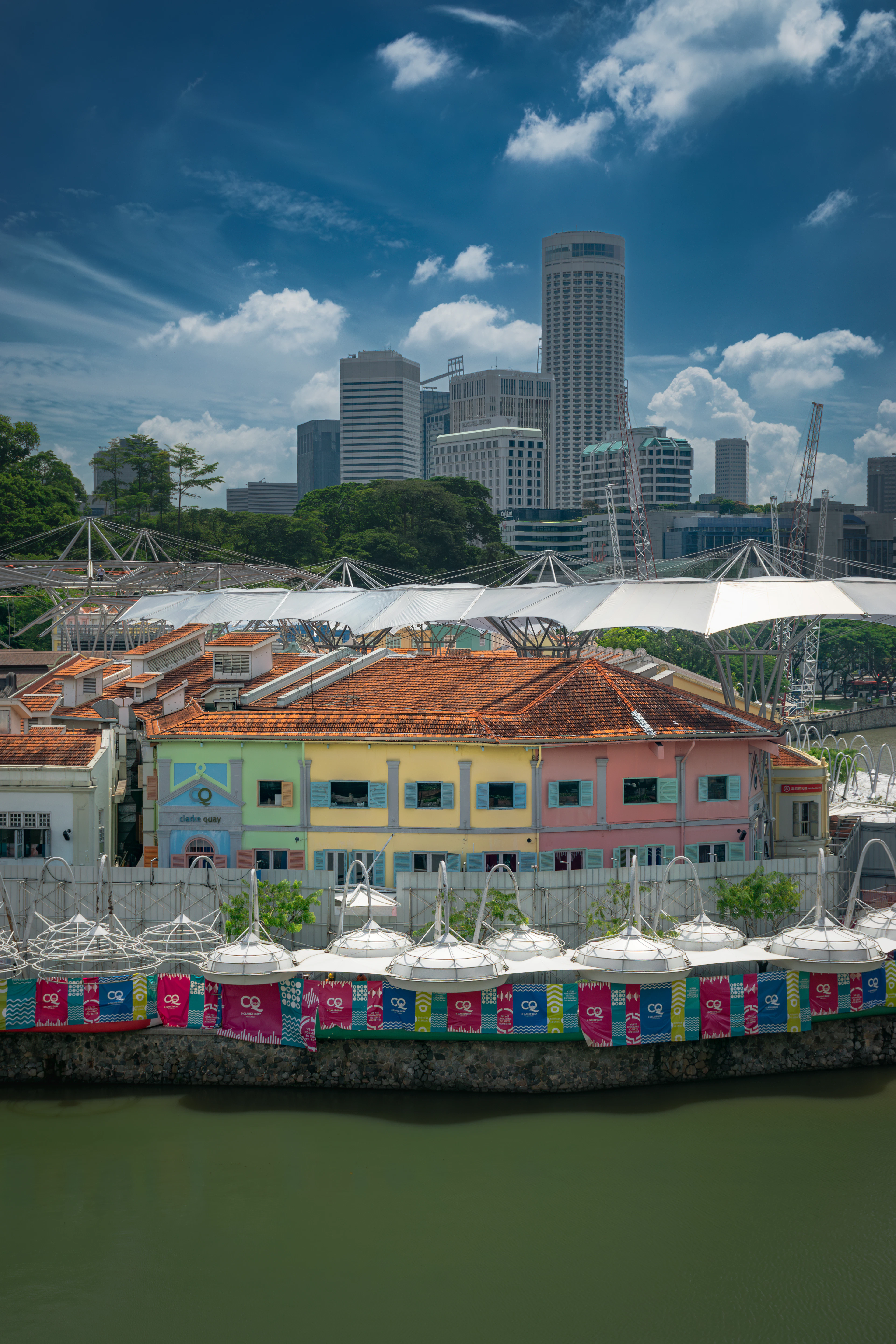 The pastel-painted warehouse shophouses and white sail canopies of Clarke Quay along the Singapore River, with the towers of the financial district rising behind under piled white cloud.