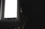 A silhouette of a person sitting contemplatively in front of a glowing window at dusk.