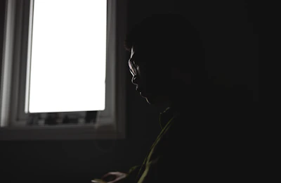 A silhouette of a person sitting contemplatively in front of a glowing window at dusk.