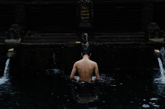 A person stands partially submerged in a dark water pool beneath a stone structure adorned with carvings and spouts. Water flows into the pool from two spouts on either side, and offerings are placed near them. The setting appears serene and contemplative.
