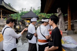 Several individuals are standing and interacting in an outdoor setting featuring traditional structures with thatched roofs. Statues and ornate architectural details are visible, and the people are dressed in a mix of traditional and modern clothing. The background includes lush green foliage.