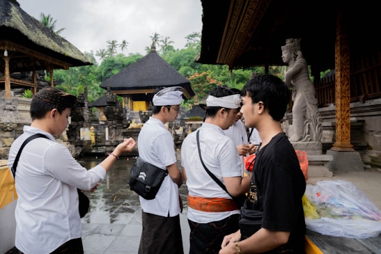 Several individuals are standing and interacting in an outdoor setting featuring traditional structures with thatched roofs. Statues and ornate architectural details are visible, and the people are dressed in a mix of traditional and modern clothing. The background includes lush green foliage.