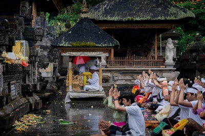 A group of people dressed in traditional attire are seated on the ground, engaging in a communal prayer or worship ceremony in front of an ornate, carved stone structure with a thatched roof. Offerings and various colorful items are placed around them.
