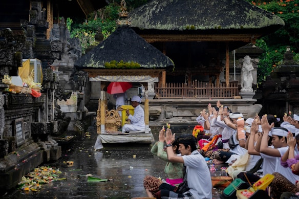 A group of people dressed in traditional attire are seated on the ground, engaging in a communal prayer or worship ceremony in front of an ornate, carved stone structure with a thatched roof. Offerings and various colorful items are placed around them.