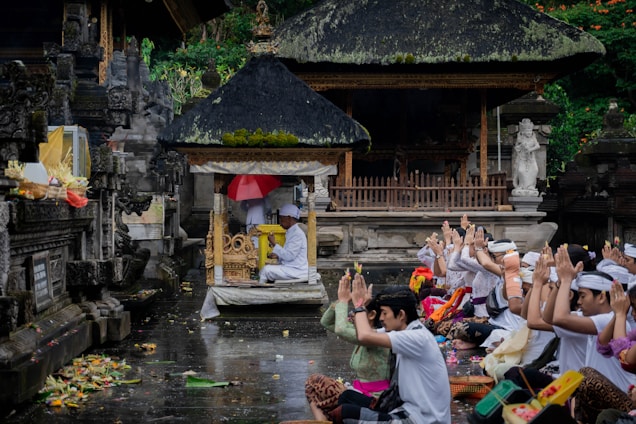 A group of people dressed in traditional attire are seated on the ground, engaging in a communal prayer or worship ceremony in front of an ornate, carved stone structure with a thatched roof. Offerings and various colorful items are placed around them.
