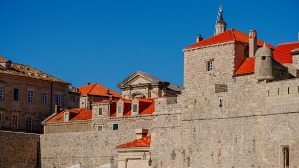 A collection of historic stone buildings with red-tiled roofs set against a clear blue sky. The architecture features tall, fortified walls and a blend of classical and medieval styles with rectangular windows and domed structures.