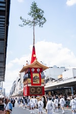 A large traditional float is being paraded down a city street, adorned with vibrant red fabric and intricate designs. It features a tall tree on top and is surrounded by people in traditional Japanese festival attire. The street is lined with spectators, and the buildings suggest an urban setting.