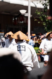 Several people are dressed in traditional attire, with wide-brimmed conical hats, gathered outdoors during an apparent cultural event or festival. The main focus is on a person wearing a white garment with a black character on the back. The atmosphere is lively with people in the background, and some greenery is present.