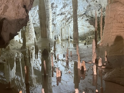 A stunning cave interior featuring numerous stalactites and stalagmites surrounded by a reflective pool. The formations are naturally ornate, creating a sense of ancient beauty and mystery. The cave walls display varied textures and colors, enhanced by subtle lighting that adds depth to the scene.