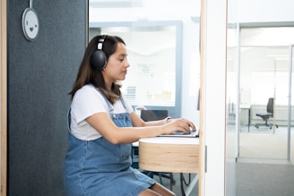A woman wearing headphones sits in a small, modern booth, working on a laptop. She is dressed in a white shirt and denim overalls. The booth has soundproof features with a smiley face ornament on the wall. In the background, there is an office space with chairs and glass partitions.