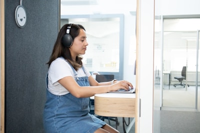 A woman wearing headphones sits in a small, modern booth, working on a laptop. She is dressed in a white shirt and denim overalls. The booth has soundproof features with a smiley face ornament on the wall. In the background, there is an office space with chairs and glass partitions.