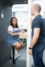 A man and a woman are engaged in conversation in a small, modern booth. The woman is seated on a stool, wearing a denim dress and white sneakers. The man, wearing a dark t-shirt and jeans, stands outside the booth with his hand resting on the door frame.