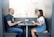Two people sitting across from each other in a modern office space, working on laptops. The setting is a small, private booth with minimal decor, featuring a partition with horizontal blinds that allow natural light to filter through. Both individuals appear engaged in conversation or collaboration.