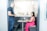A man in a dark t-shirt is standing and talking to a woman seated at a small desk in a modern office setting. The woman is wearing a pink patterned top and a bright pink skirt, and she is using a laptop. The area is enclosed with glass partitions, offering a semi-private workspace.