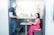 A man in a dark t-shirt is standing and talking to a woman seated at a small desk in a modern office setting. The woman is wearing a pink patterned top and a bright pink skirt, and she is using a laptop. The area is enclosed with glass partitions, offering a semi-private workspace.