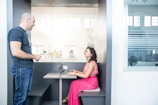 A man and a woman are having a conversation in a modern office setting. The woman is seated at a small booth with a laptop in front of her, while the man stands beside the booth, talking to her. The room has large windows and glass partitions that create a bright and open atmosphere.