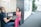 Two people are seated at a small gray table having a conversation in a modern, bright office space. One person is using a laptop, while the other is smiling, wearing a pink floral top and pink skirt. The area is well-lit with natural light, and there are large green plants and framed pictures along the walls.