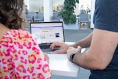 Two people are interacting with a laptop on a white standing desk. The laptop screen displays a webpage related to social wall solutions for hashtag campaigns. The setting appears to be a modern office with several chairs, tables, and a large plant in the background. One person is wearing a colorful floral top and the other is in a dark t-shirt.