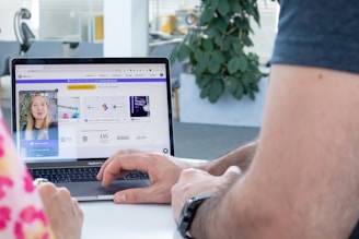 A person is interacting with a laptop displaying a website on the screen. The site features a video call window with a woman and several graphic elements such as logos and text. The environment appears to be an office setting with a plant visible in the background.