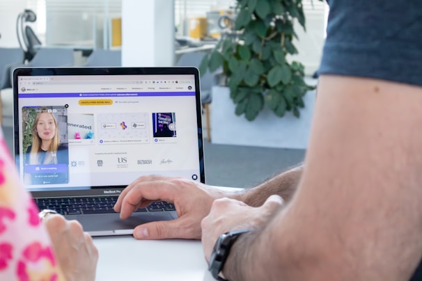 A person is interacting with a laptop displaying a website on the screen. The site features a video call window with a woman and several graphic elements such as logos and text. The environment appears to be an office setting with a plant visible in the background.