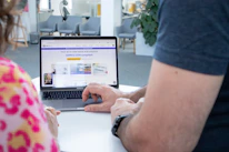 Two people are seated in front of a laptop screen that displays a webpage. The webpage features text about a social wall solution compliant with GDPR and CCPA. Both of the individuals' hands are visible as they interact with the laptop. The background shows a modern office environment with chairs and a potted plant.