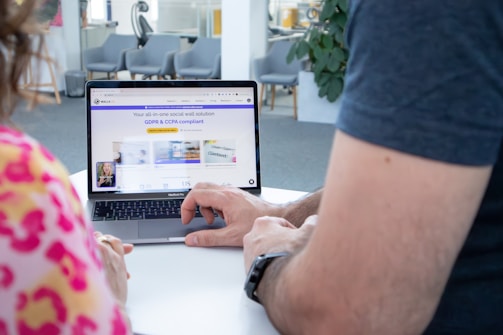 Two people are seated in front of a laptop screen that displays a webpage. The webpage features text about a social wall solution compliant with GDPR and CCPA. Both of the individuals' hands are visible as they interact with the laptop. The background shows a modern office environment with chairs and a potted plant.