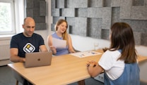 Three individuals are seated around a rectangular wooden table in a modern office setting. One person is working on a laptop, another is holding and reading from a paper, and the third person is attentively listening. The walls are decorated with grey acoustic panels and there is a window letting natural light in.