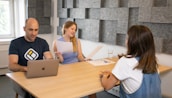 Three people are seated around a wooden table in an office setting with grey acoustic panels on the walls. One person is using a laptop, another holds a piece of paper, and the third is listening attentively. A glass of water is also visible on the table.