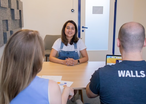 A woman with long hair is sitting at a wooden table, facing two people. She appears to be in conversation or an interview. One person has blonde hair and is wearing a blue top, while the other is bald and wearing a dark shirt with 'WALLS' written on the back. A laptop with a yellow screen is open in front of the bald person. The room has a minimalist design with a chair and a textured panel on the wall.