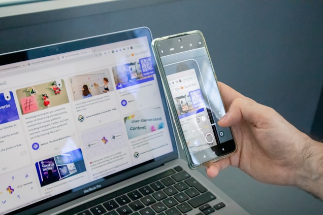 A person holding a smartphone in their hand, capturing a photograph of a laptop screen. The laptop displays a webpage with various social media content and images. The smartphone screen shows the camera interface with the laptop screen content visible.