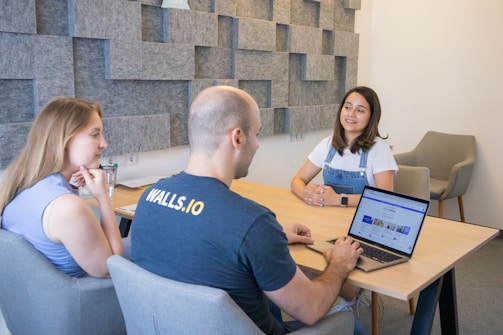 Three people are gathered in a modern office setting. Two women and one man are seated around a rectangular wooden table. The man, with a laptop open in front of him, is typing. The woman on the right, wearing a white t-shirt and denim overalls, is smiling. The atmosphere appears relaxed and professional. The wall behind them is covered with geometric acoustic panels in shades of gray.