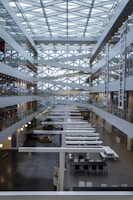 A modern interior of a multi-story atrium featuring a geometric glass ceiling with crisscrossing beams. The space is lined with glass-walled offices and corridors on both sides. Long white tables and chairs populate the ground floor, and there are various banners with national flags along the walls.