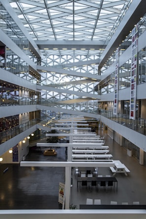 A modern interior of a multi-story atrium featuring a geometric glass ceiling with crisscrossing beams. The space is lined with glass-walled offices and corridors on both sides. Long white tables and chairs populate the ground floor, and there are various banners with national flags along the walls.