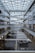 A modern interior of a multi-story atrium featuring a geometric glass ceiling with crisscrossing beams. The space is lined with glass-walled offices and corridors on both sides. Long white tables and chairs populate the ground floor, and there are various banners with national flags along the walls.