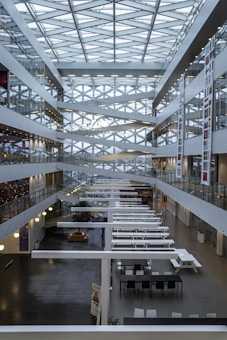A modern interior of a multi-story atrium featuring a geometric glass ceiling with crisscrossing beams. The space is lined with glass-walled offices and corridors on both sides. Long white tables and chairs populate the ground floor, and there are various banners with national flags along the walls.