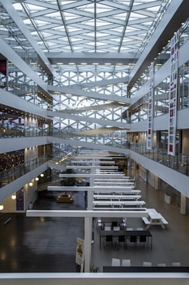 A modern interior of a multi-story atrium featuring a geometric glass ceiling with crisscrossing beams. The space is lined with glass-walled offices and corridors on both sides. Long white tables and chairs populate the ground floor, and there are various banners with national flags along the walls.