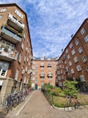 A courtyard surrounded by brick apartment buildings with balconies adorned with plants and flowers. Bicycles are lined up along the left side, and a well-kept garden with neatly trimmed bushes and blooming flowers is situated near the center. The sky overhead is partly cloudy, adding a touch of depth to the scene.
