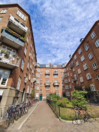 A courtyard surrounded by brick apartment buildings with balconies adorned with plants and flowers. Bicycles are lined up along the left side, and a well-kept garden with neatly trimmed bushes and blooming flowers is situated near the center. The sky overhead is partly cloudy, adding a touch of depth to the scene.