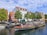 A canal tour boat filled with people is traveling on a calm waterway, surrounded by parked smaller boats. Colorful, traditional-style buildings with red and white facades line the street, accompanied by green trees. The sky is partly cloudy, adding to the picturesque scene.