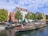 A canal tour boat filled with people is traveling on a calm waterway, surrounded by parked smaller boats. Colorful, traditional-style buildings with red and white facades line the street, accompanied by green trees. The sky is partly cloudy, adding to the picturesque scene.