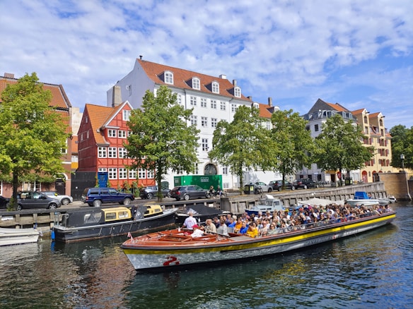 A canal tour boat filled with people is traveling on a calm waterway, surrounded by parked smaller boats. Colorful, traditional-style buildings with red and white facades line the street, accompanied by green trees. The sky is partly cloudy, adding to the picturesque scene.