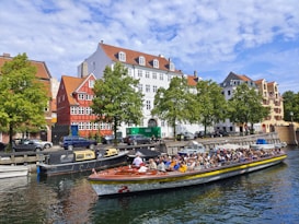 A canal tour boat filled with people is traveling on a calm waterway, surrounded by parked smaller boats. Colorful, traditional-style buildings with red and white facades line the street, accompanied by green trees. The sky is partly cloudy, adding to the picturesque scene.