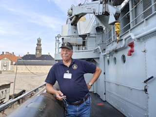 A man in a navy blue polo shirt and cap is standing on a ship's deck with an old building in the background. He has a name tag on his shirt and is holding a pair of glasses in one hand. The ship has various metallic structures and equipment visible, including a crane and pipes.
