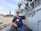 A man in a navy blue polo shirt and cap is standing on a ship's deck with an old building in the background. He has a name tag on his shirt and is holding a pair of glasses in one hand. The ship has various metallic structures and equipment visible, including a crane and pipes.