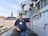 A man in a navy blue polo shirt and cap is standing on a ship's deck with an old building in the background. He has a name tag on his shirt and is holding a pair of glasses in one hand. The ship has various metallic structures and equipment visible, including a crane and pipes.