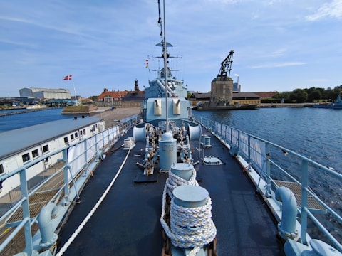 A large warship is moored at a harbor near industrial buildings and clear blue skies. The deck of the ship is equipped with various machinery, ropes, and railings. A flag waves on the left side, while the right shows a crane structure and water body.