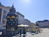 An ornate kiosk with a decorative roof, surrounded by a cobblestone plaza. The kiosk offers services like Segway tours and draft beer to go. Outdoor seating with tables and umbrellas is visible, alongside a street lined with historical buildings under a clear blue sky.