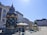 An ornate kiosk with a decorative roof, surrounded by a cobblestone plaza. The kiosk offers services like Segway tours and draft beer to go. Outdoor seating with tables and umbrellas is visible, alongside a street lined with historical buildings under a clear blue sky.
