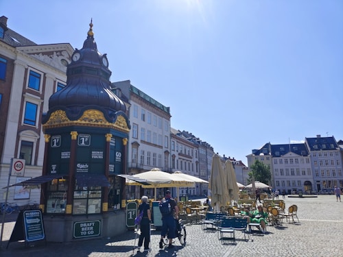 An ornate kiosk with a decorative roof, surrounded by a cobblestone plaza. The kiosk offers services like Segway tours and draft beer to go. Outdoor seating with tables and umbrellas is visible, alongside a street lined with historical buildings under a clear blue sky.