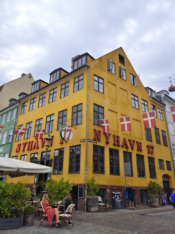A vibrant yellow building featuring prominent red signage 'Nyhavn 17' and adorned with Danish flags. The building has black-framed windows and a small outdoor seating area with people sitting at tables. Potted plants add green to the scene, and the street is cobblestoned, giving a quaint and historic atmosphere.